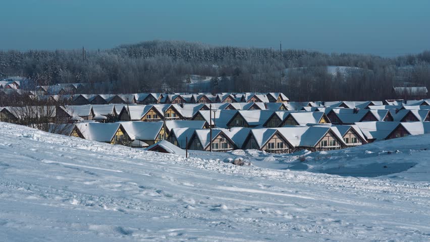 A winter landscape in a village, with mountains and a forest in the background and the roofs of houses with chimneys covered in snow in the front