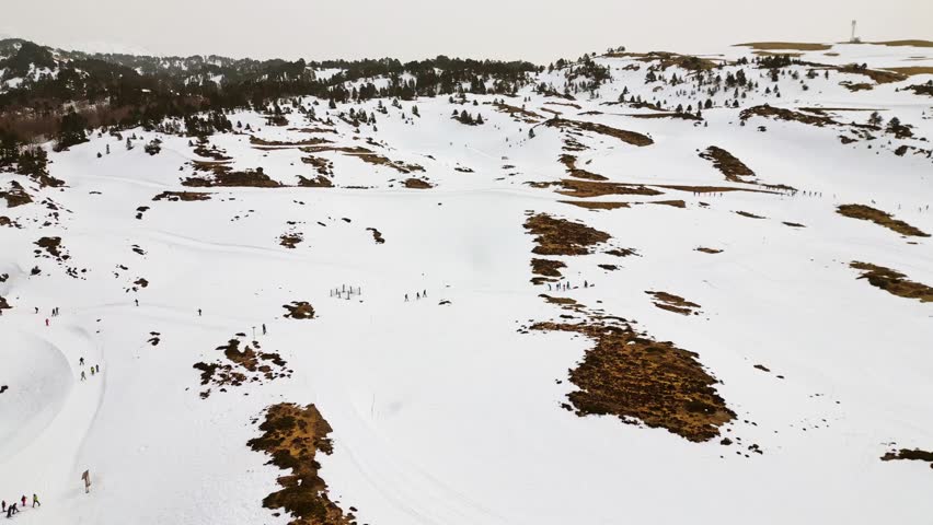Aerial drone view flying backwards over a vast snowy mountain landscape, revealing groups of people practicing winter sports like snowshoeing and hiking across the white hills and pine forests
