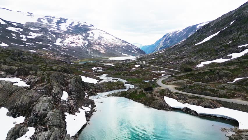 Aerial view of the winding Old Strynefjell Mountain Road through a rugged landscape with snow-capped peaks, glacial streams, and rocky terrain in Stryn Municipality, Vestland, Norway.