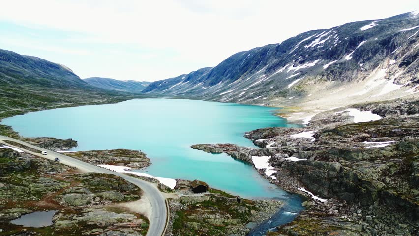 Aerial view of the turquoise glacial lake and the winding Old Strynefjell Mountain Road surrounded by rocky mountain terrain in Stryn Municipality, Vestland, Norway.