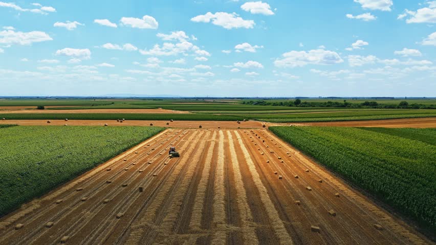 Aerial view of a tractor baling hay in a large field with rows of hay bales and surrounding green agricultural crops under a blue sky in Jazak, Vojvodina, Serbia.