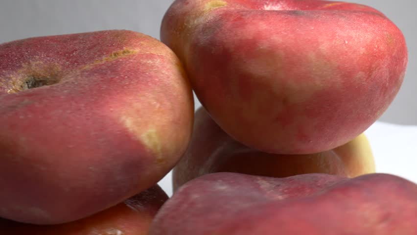 Close-up of fresh flat Saturn peaches rotating on a white plate, showcasing their fuzzy red skin and unique donut shape under bright studio lighting.