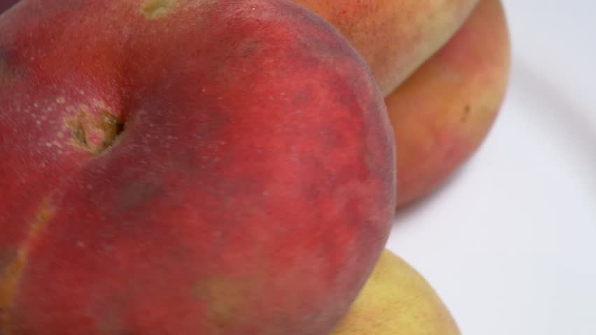 Close-up of fresh flat Saturn peaches rotating on a white plate, showcasing their fuzzy red skin and unique donut shape under bright studio lighting.