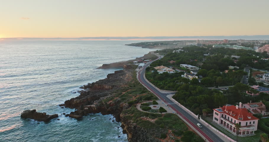Travel to Portugal. Cascais coastline revealing a winding road, luxury villas, dense vegetation along rocky shore, with gentle Atlantic Ocean waves during a serene sunset. Aerial view drone panorama