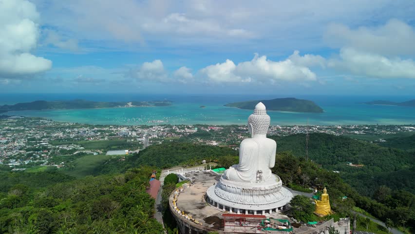 Aerial view of the white marble Big Buddha statue set atop a lush green hill overlooking the coastal landscape and blue sea in Phuket, Thailand.