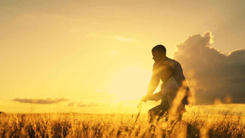 Dad helps child son ride bike. Dad teaches child wearing safety helmet to ride bicycle Family in meadow. Child rides bicycle. Kid dad play together, sunset. Child boy dream learns to ride bicycle, sun