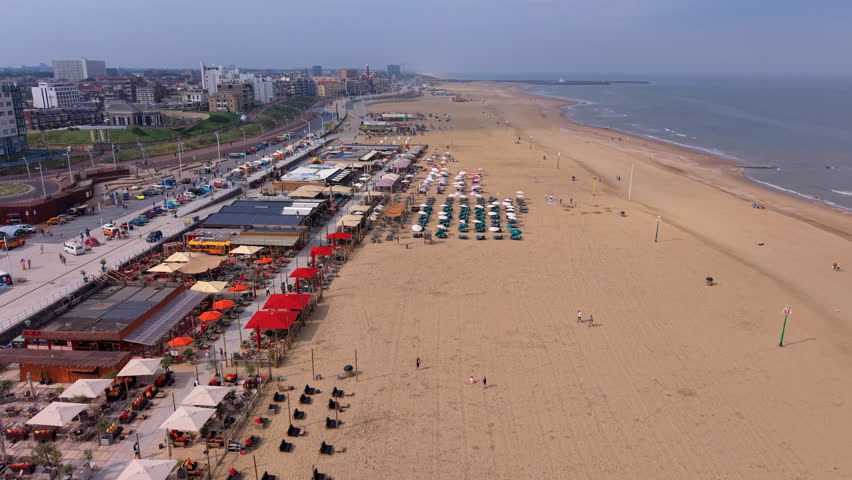 A magnificent wide-angle aerial photograph capturing the bustling coastal infrastructure and leisure layout of Scheveningen beach. The left foreground is filled with several large beachfront restaurant and cafe complexes featuring spacious outdoor wooden terraces decorated with striking bright red and orange parasols. Adjacent to the venues, a wide paved promenade and parallel roadways are lined with parked vintage cars, camper vans, and light municipal traffic next to the rising urban skyline of the resort town. On the sandy shore, orderly geometric rows of beach umbrellas and sun loungers are arranged for visitors. The vast sandy beach stretches far into the background, meeting the calm waters of the North Sea on the right under bright morning summer daylight. This high-altitude overview perfectly documents maritime tourism, seaside hospitality design, and regional urban planning.