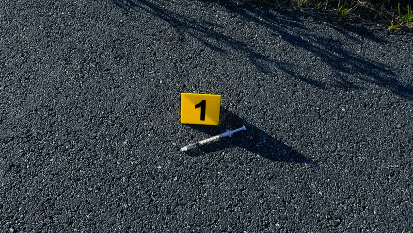 Yellow evidence marker number one placed next to a used syringe on the asphalt, marking a piece of evidence at an outdoor crime scene investigation related to drug abuse or overdose
