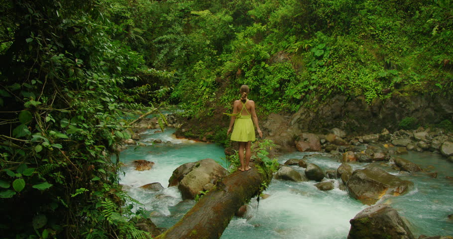 Traveler woman stands atop a fallen log over the turquoise river in Blue Falls reserve Costa Rica