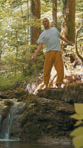A man practices yoga on a moss-covered cliff beside a cascading waterfall in a lush forest. His focused pose exudes balance and tranquility, embodying a zen-like state amidst nature