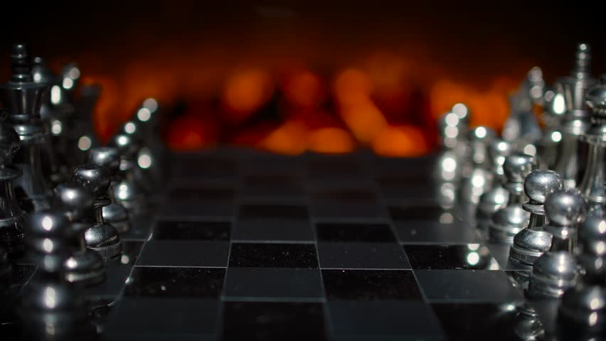 A close-up of silver chess pieces on a board with dramatic orange fire-like bokeh lighting creating a moody atmospheric scene.