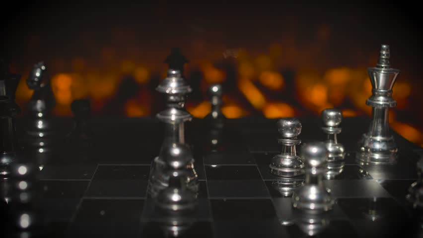 A hand reaching to move a silver chess piece on a board during a game with dramatic orange fire-lit background.