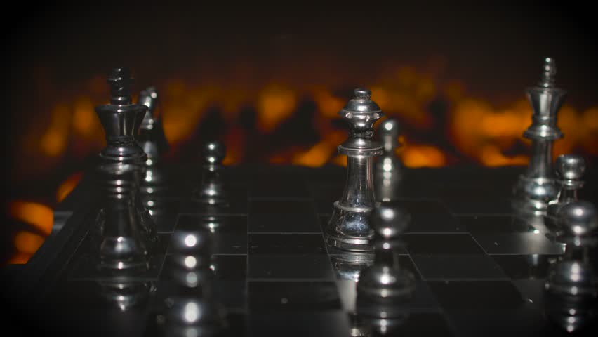 A hand knocking over or moving a silver chess piece on a board with dramatic orange lighting and scattered pieces visible.