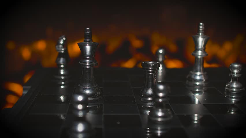 A close-up of silver chess pieces on a board with the king piece prominent against a dramatic orange fire-lit background.