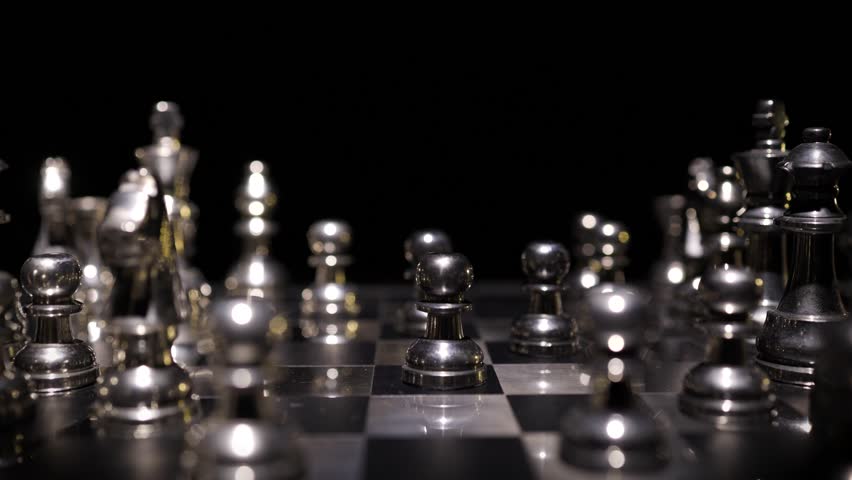 A hand moving or placing a silver chess piece on a board with other pieces in dramatic contrast against a dark background.