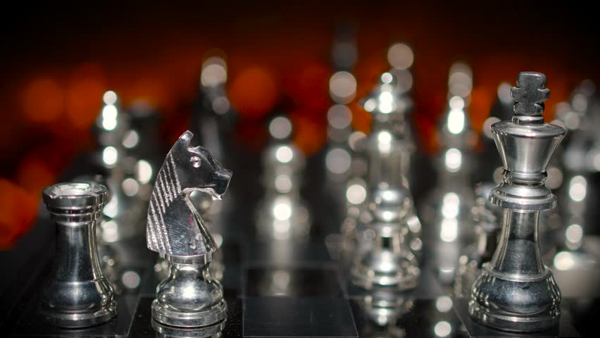Silver chess pieces arranged on a board with dramatic dark atmospheric lighting and selective focus highlighting key pieces.