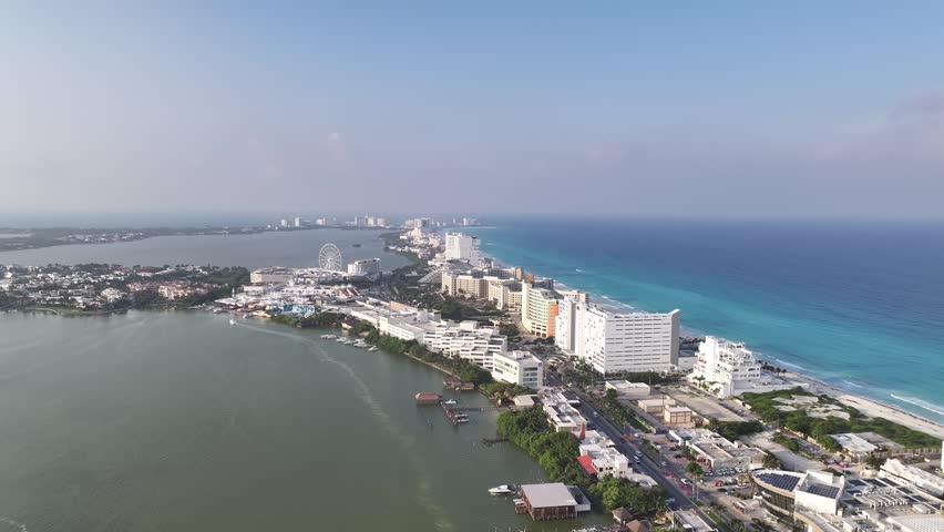 Aerial view of the Cancun hotel zone, featuring high-rise resorts, the Nichupte Lagoon, the Caribbean Sea, and a Ferris wheel under a clear sky in Cancun, Quintana Roo, Mexico.