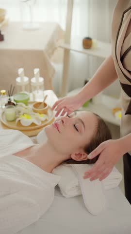 Beautiful woman getting head and facial massage treatment at beauty spa. Attractive girl lying down on massage bed, getting relaxing physiotherapy service from masseuse for body care in spa clinic.