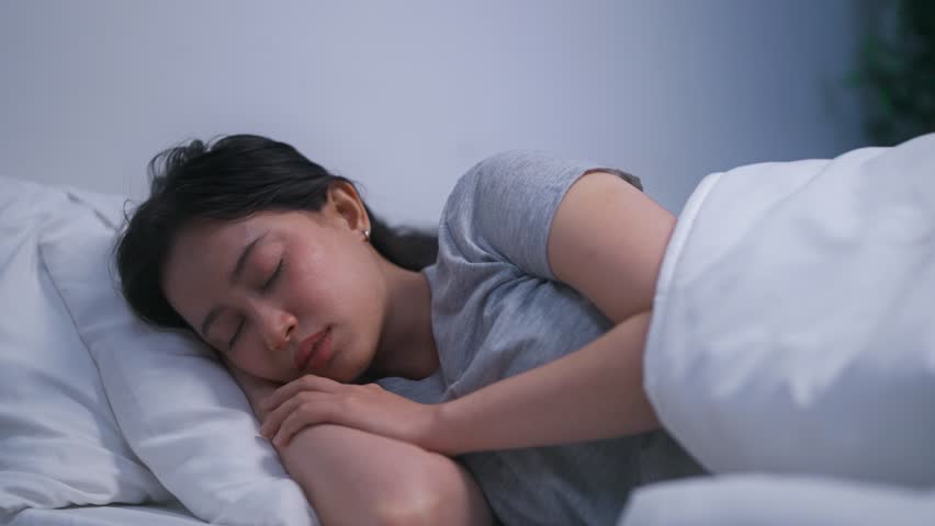 Asian woman struggling with insomnia and having a nightmare at night. Attractive young girl eyes closed, lying down on bed in bedroom, showing distress and discomfort while battles a bad dream.