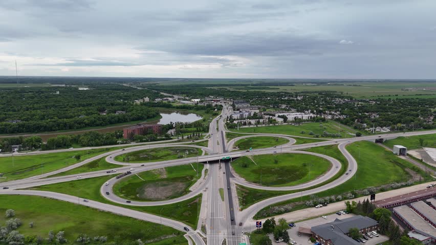 Static drone high above the Pembina Highway and Perimeter Highway cloverleaf interchange in south Winnipeg, looking toward St. Norbert with traffic, the Red River, green grass and partly cloudy summer