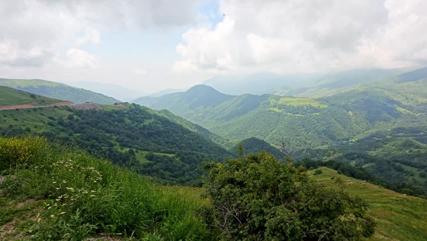 Rolling green hills stretch under cloudy skies. Lush vegetation covers slopes and valleys