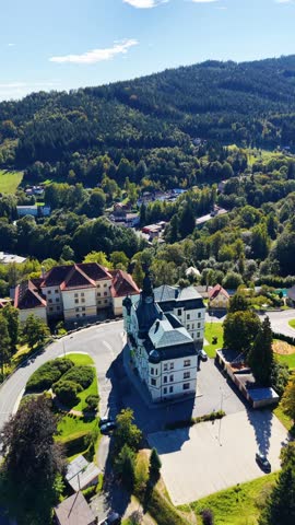 Aerial view of the historic Tanvald Town Hall surrounded by lush green forests and residential buildings in the town of Tanvald, Liberec Region, Czechia.