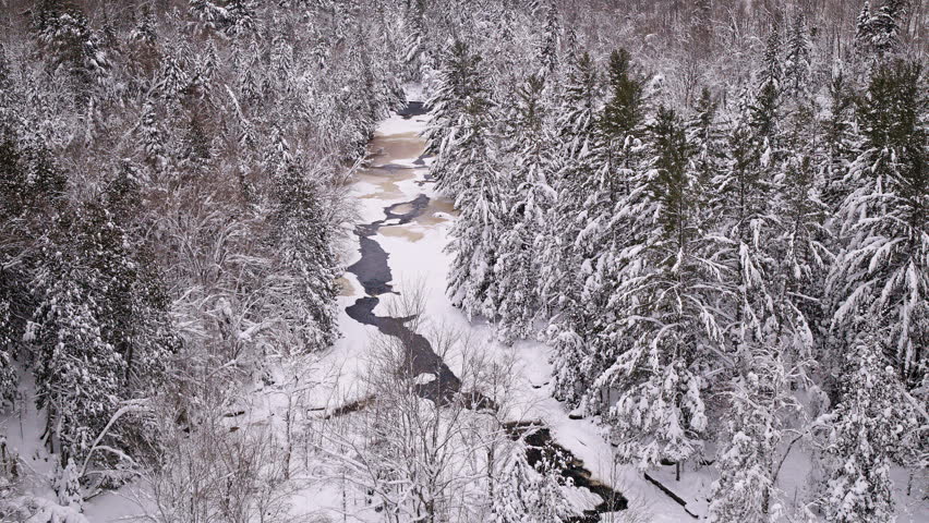 Sweeping drone shot of a partially frozen river surrounded by untouched snowy forest in Northern Michigan.