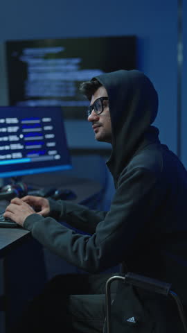 Young man in black hooded sweatshirt and glasses typing at desk with data screens in dark room, cyber crime and hacking concept.