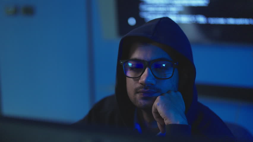 Close up of young man in black hood with eyeglasses resting chin on hand, planning cyber attack in dark room, hacking and cyber crime concept.