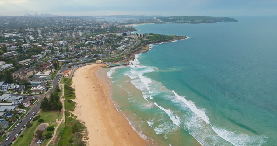 Amazing Manly Beach sandy shore with waves breaking on the aqua ocean. The coastal road leading to a peninsula featuring residential buildings and distant Sydney city skyline under a cloudy sky