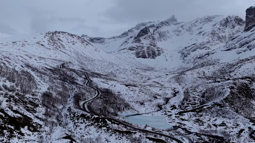 Aerial view of a winding road through snowy mountains near Rekvik with a frozen lake under an overcast sky in Tromso, Troms, Norway.