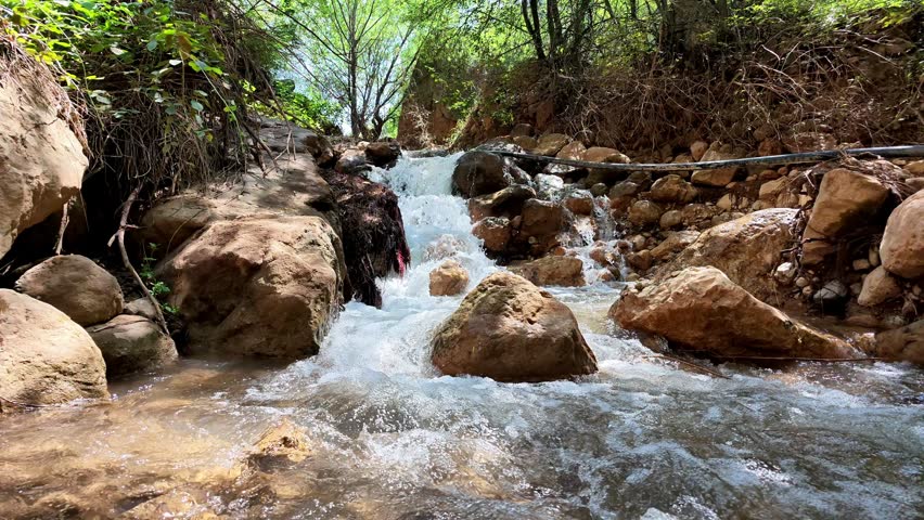 Clear mountain stream flows over smooth stones. Water cascades down rocky terrain