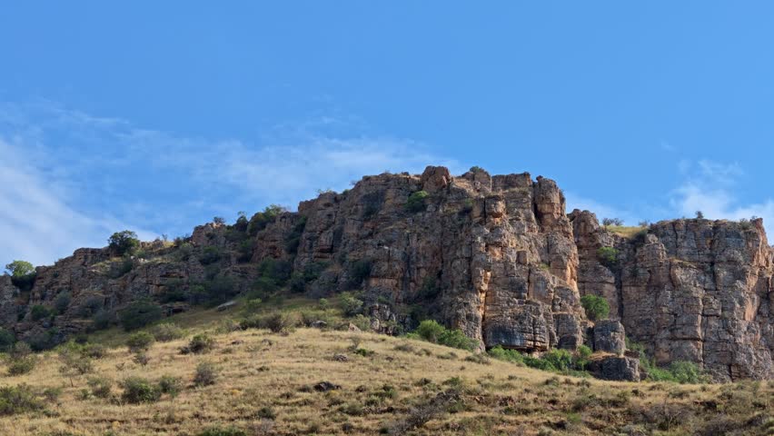 Rugged rock formations rise under clear blue sky. Dry grass covers foreground slopes