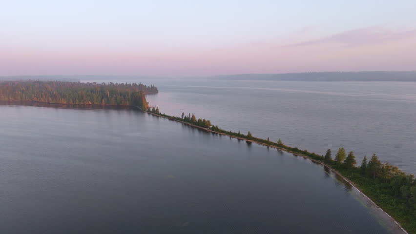Drone pullback revealing a narrow sandy peninsula extending into Lake Nagagamisis beneath a soft pink morning sky with light haze drifting over the remote Northern Ontario landscape.