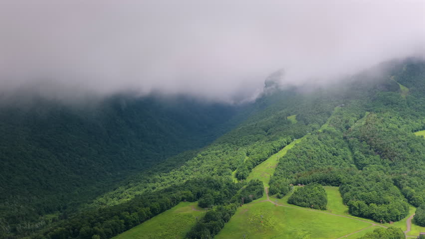 Aerial shot of sunlight breaking through clouds onto the green summer slopes of Stowe Mountain Resort in Vermont, with fog partially obscuring Smuggler