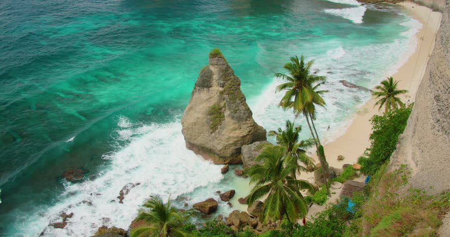 Jagged limestone rock stands in turquoise ocean water near sandy beach with palm trees