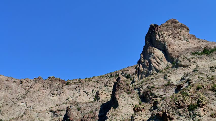 Rugged mountain peak rises sharply against clear blue sky. Rock formations show erosion patterns