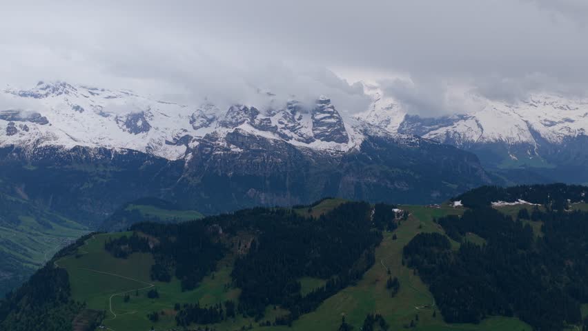 Aerial view of Stanserhorn Cabrio with snow-capped mountain peaks and lush green alpine slopes under a cloudy sky in Stans, Nidwalden, Switzerland.
