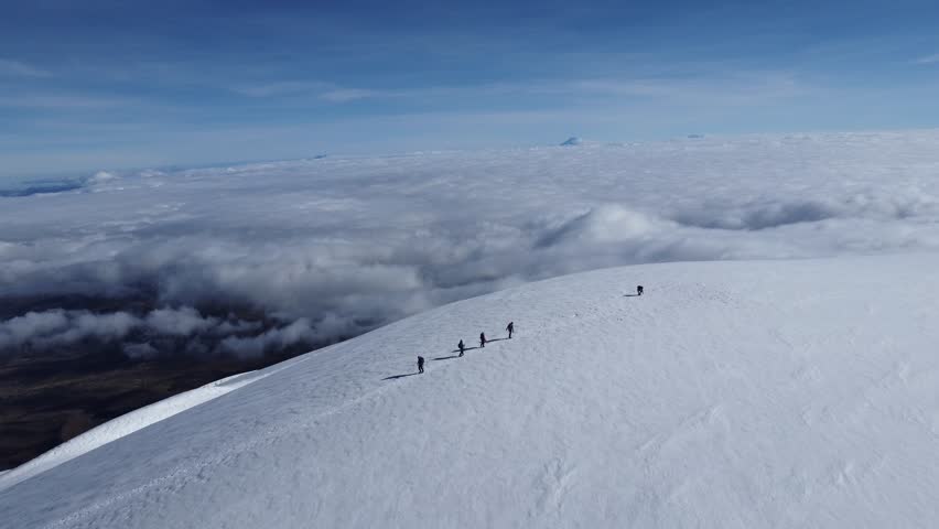 Five people trekking on a snowy mountain slope with a vast cloudscape in the background under a blue sky, viewed from a high vantage point.