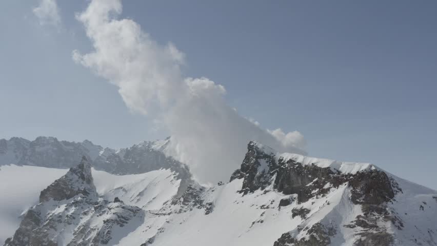 Snow-covered mountain peaks with rocky ridges and a plume of steam or cloud rising against a clear blue sky in a winter landscape.