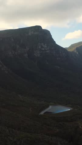 Steep mountain landscape with cliffs, shadows, and a small reflective lake in the valley below, under a partly cloudy sky with sunlight on distant slopes.