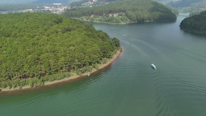 Drone footage captured above Tuyen Lam Lake in Da Lat, Lam Dong, Vietnam. The drone gradually descends toward a pine forest while following a small boat moving across the calm green water. Light mist hangs over the landscape, creating a peaceful and atmospheric scene that highlights the natural beauty of the lake and surrounding highlands.
