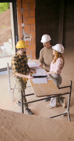 Adult couple with hard hat discussing building plans with a contractor at a house construction site.