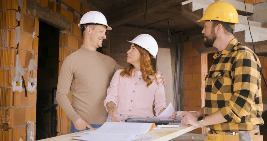 Adult couple with hard hat discussing building plans with a contractor at a house construction site.