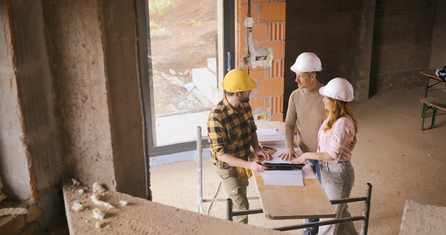 Adult couple with hard hat discussing building plans with a contractor at a house construction site.