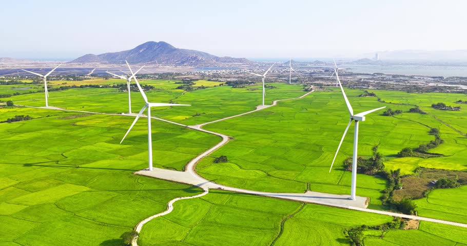 Bright sunny day at the Ninh Thuan wind farm, Vietnam, featuring modern wind turbines rising above vast green agricultural fields. The clear blue sky, winding rural roads, and renewable energy infrastructure create a striking landscape of sustainability and natural beauty.