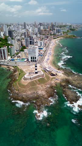 Aerial view of Farol da Barra lighthouse on the rocky coastline of Salvador, with the urban skyline, sandy beach, and turquoise Atlantic Ocean waters in Salvador, Bahia, Brazil.