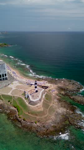Aerial view of the historic Farol da Barra lighthouse situated on a rocky point with sandy beaches, turquoise ocean waters, and urban buildings in Salvador, State of Bahia, Brazil.