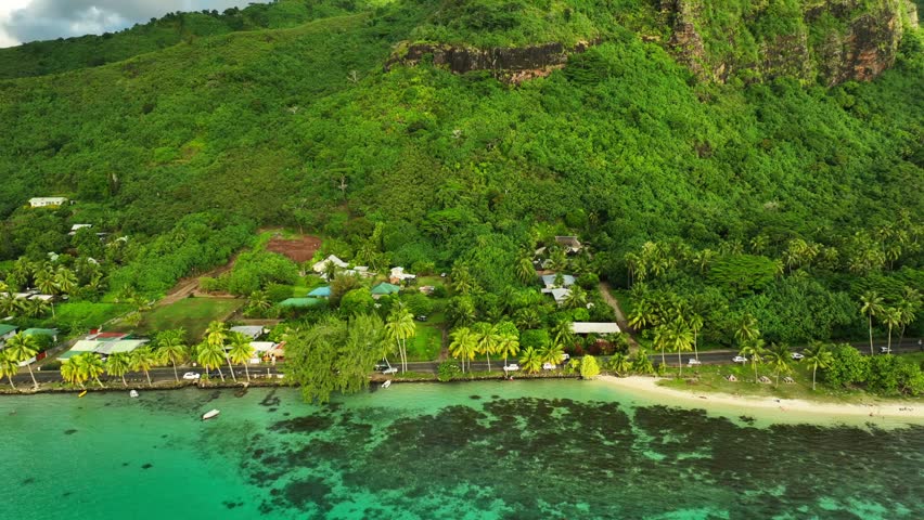 Village homes line Ta’ahiamanu Beach shoreline, Moorea’s coral reef and turquoise lagoon stretch beneath green mountain slopes within French Polynesia’s Society Islands. Aerial drone approaching