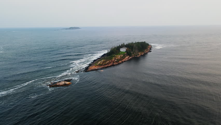 Sunset aerial of a secluded coastal mansion on a private island near Popham Beach, Maine, with surrounding evergreen forest and rocky coastline in the middle of a calm blue Atlantic Ocean.